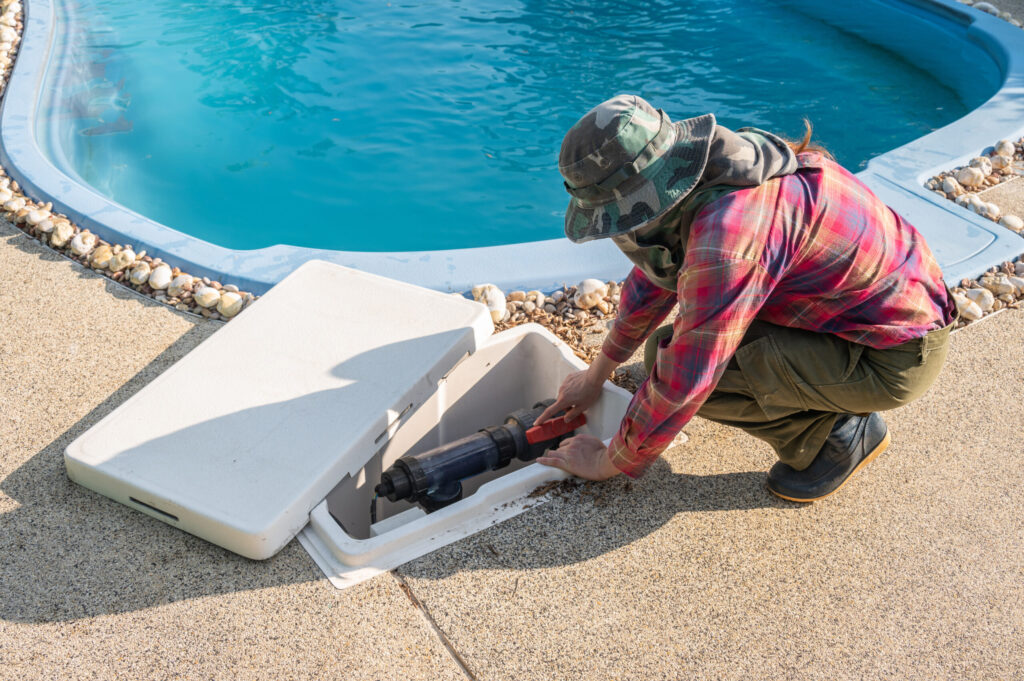 AdobeStock_615631240-scaled-1jpeg - Wrigley's Pool Co. - Wrigley's Pool Co. Pool maintenance worker crouching by a bright blue pool, using a handheld tool on a white cooler-like equipment box near the edge of the deck.