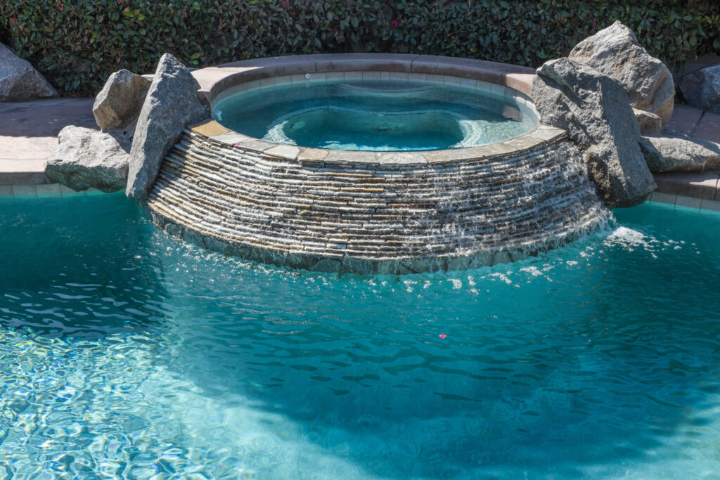 AdobeStock_89391793-scaled-1jpeg - Wrigley's Pool Co. - Wrigley's Pool Co. Circular stacked-stone fountain in a turquoise pool, with large rocks flanking the structure and a leafy hedge in the background