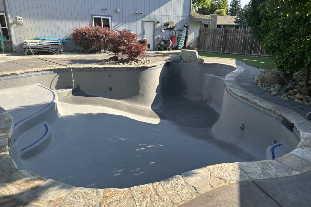 Pool-Drain-jpg - Wrigley's Pool Co. - Wrigley's Pool Co. Empty in-ground backyard pool with gray plaster, stone coping, and surrounding patio stones; outdoor shed and landscaping visible in the background.